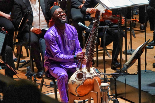 A musician in a purple outfit plays the kora on stage at Ulster Hall with an orchestra; he is seated and appears to be singing or smiling broadly, creating a boundary breaking performance.