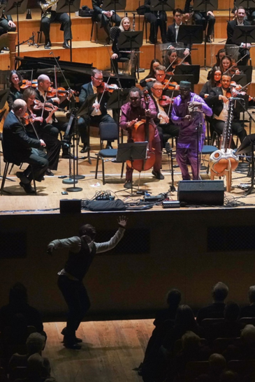 A musician in purple plays in front of an orchestra on stage at Ulster Hall while another performer dances energetically near the audience, creating a boundary-breaking concert experience.