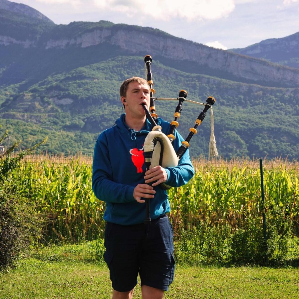 A man stands outdoors playing bagpipes in front of a field, with mountains and trees in the background, capturing a serene moment in pictures reminiscent of the Festival Berlioz.