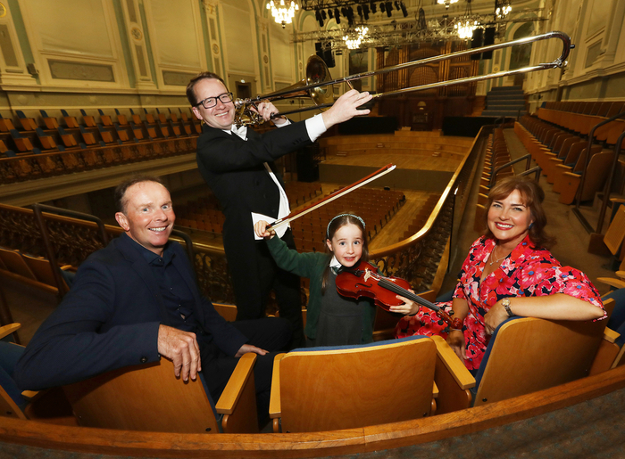 Four people sit in theater seats; a man in a tuxedo stands and plays a large trombone, while a girl, as part of the Ulster Orchestra Schools Concerts programme, holds a violin and bow, accompanied by a smiling man and woman nearby.