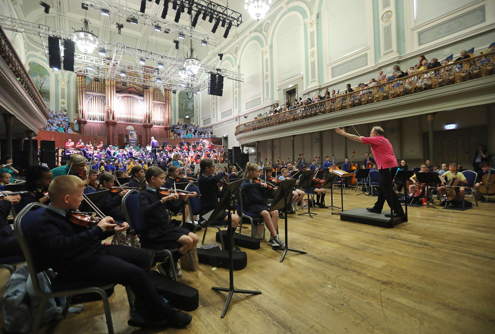 A conductor leads a youth orchestra and choir during a performance in a large, ornate concert hall with a seated audience, highlighting the musical achievements showcased in their 2023 review.