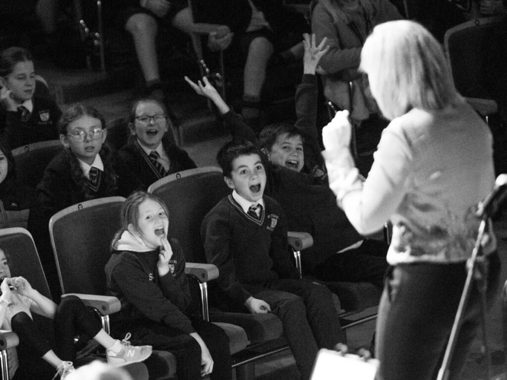 A group of schoolchildren in uniforms sit in theater seats, some raising their hands and reacting energetically, while a woman stands in front addressing them during a lively 2023 review session.