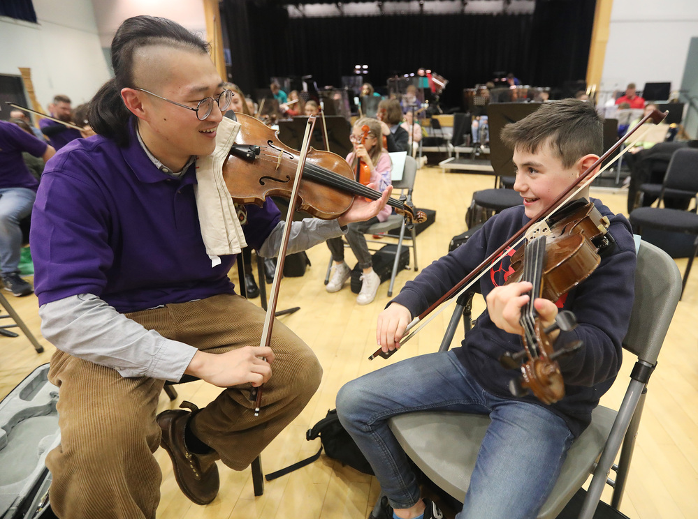 An adult and a child sit side by side in a music hall, both holding violins and playing together while smiling—a heartwarming moment from 2023 in Review. Other musicians are visible in the background.