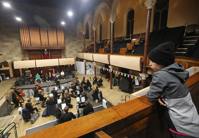 A person in a beanie watches an orchestra rehearsal from a balcony inside a large, high-ceilinged hall—a reflective moment for 2023 in Review.
