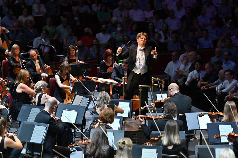 Daniele Rustioni leads the Ulster Orchestra during a performance on stage, with musicians playing string instruments and an audience seated in the backgroundโa true retrospective of orchestral artistry.