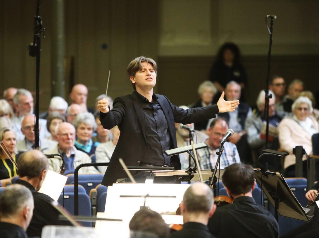 Daniele Rustioni leads the Ulster Orchestra during a performance, with musicians playing passionately as the audience enjoys the music in the background.