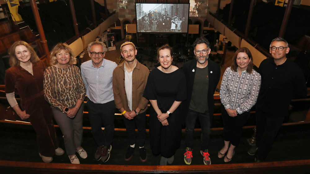 Eight people stand in a row on Townsend Street, posing for a group photo indoors with a projected black-and-white image and Sound Links visible in the background.