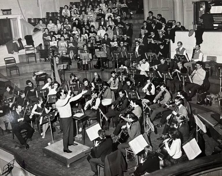 A conductor, reminiscent of Sergiu Comissiona in 1969, leads an orchestra and choir during a rehearsal in a concert hall. Musicians with instruments and choir members are seated on tiered benches behind them for a UO Playback session.