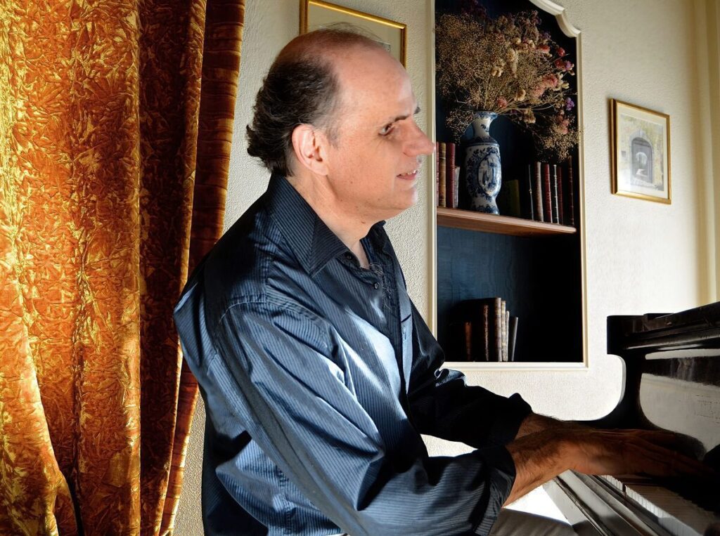A man in a dark shirt plays the piano next to gold curtains, with framed pictures and a vase of dried flowers on a shelf behind him, evoking the elegance of Viennaโs Musikverein Golden Hall in 1986.