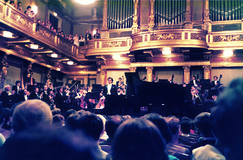 A pianist performs on a grand piano with an orchestra in the Musikverein's Golden Hall as an audience watches, evoking the elegance of 1986.