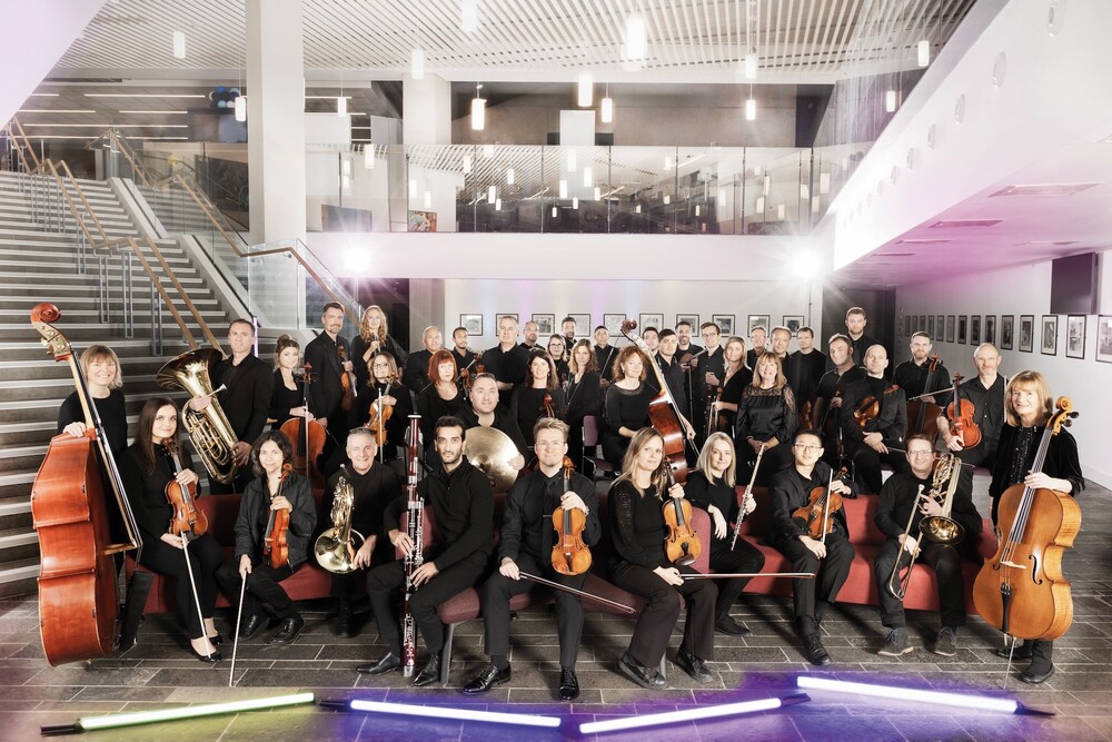 A large orchestra group poses with their instruments in a modern indoor setting, arranged on steps and seats, with string, brass, and woodwind sections visible.