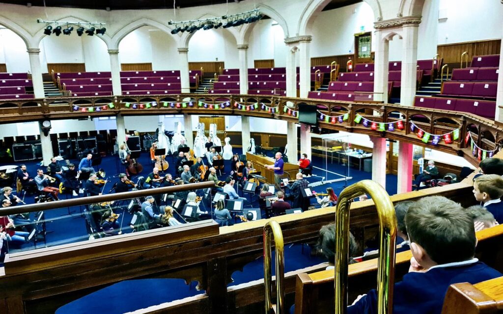 Musicians rehearse The Class(ic) Symphony with an orchestra in a grand hall with arched architecture, as audience members, including children, watch from the balcony.