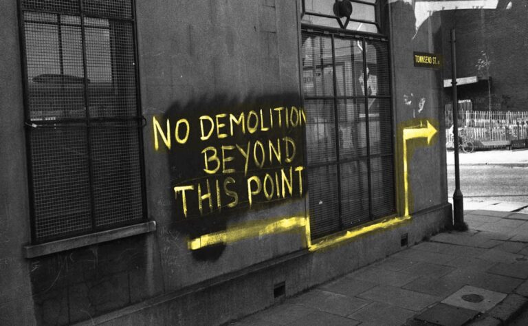 A brick building wall with yellow graffiti reading "NO DEMOLITION BEYOND THIS POINT" and a yellow arrow pointing right, creating a striking scene that feels like a bold celebration of boundaries.