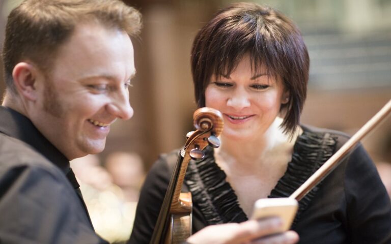 A man and a woman, holding a violin and bow, stand closely together and look at a smartphone, smiling in a well-lit indoor setting—ready for their Let's Play at Home moment.