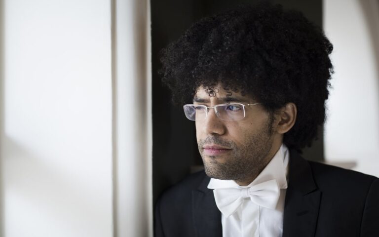 Rafael, a man with curly hair and glasses in a tuxedo and white bow tie, stands indoors, looking thoughtfully out of the window on the Final Derry~Londonderry Date.
