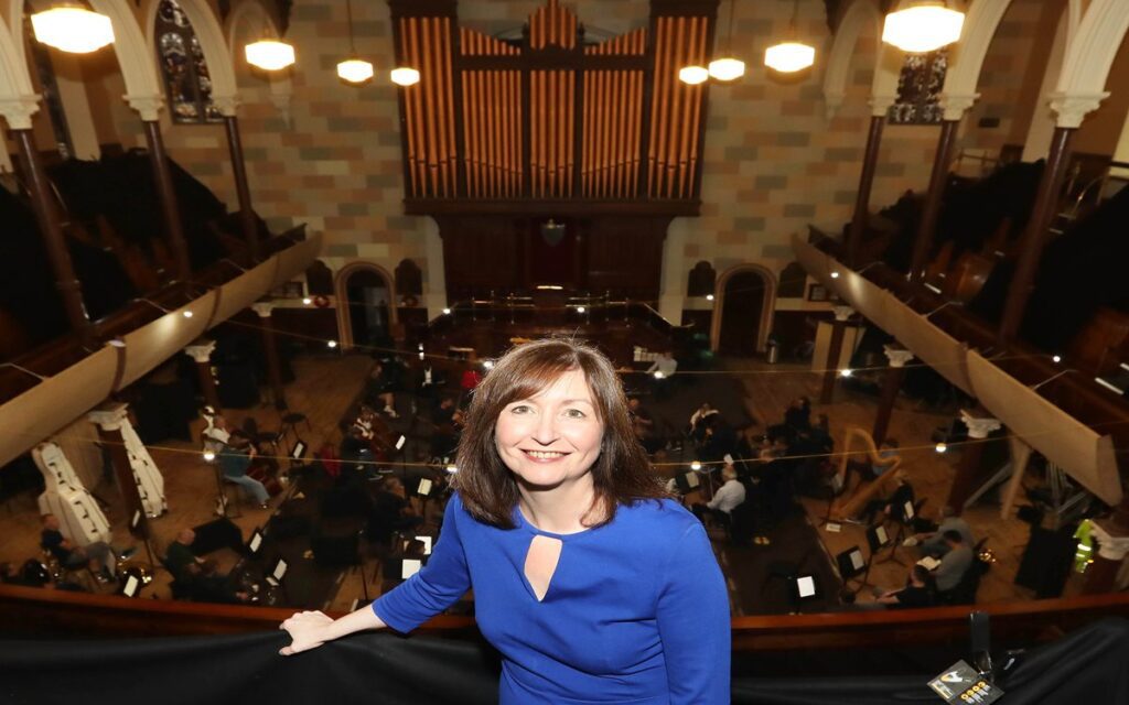 A woman in a blue dress stands on a balcony overlooking the Ulster Orchestra rehearsal in a church with high ceilings and a large pipe organ, embodying the poise of Chief Executive Auveen Sands.