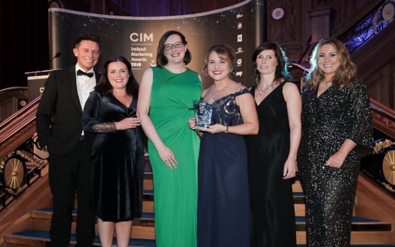 Six people from the Marketing Team pose on a staircase at the CIM Ireland Marketing Awards 2018, with one woman holding a trophy, celebrating Awards Success.