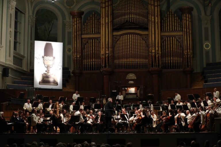 The Ulster Orchestra performs on stage in front of a grand pipe organ, with a conductor leading, as the BBC Radio 3 Afternoon Concert and a vintage BBC microphone image are projected on a screen.