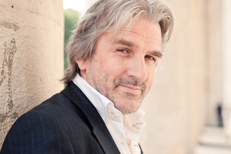A middle-aged man with gray hair and a beard, wearing a dark blazer and white shirt, stands outdoors leaning against a light stone wall, looking at the camera with a slight smile, as if ready to attend Rachmaninov Piano Concerto No.2 at Ulster Hall.
