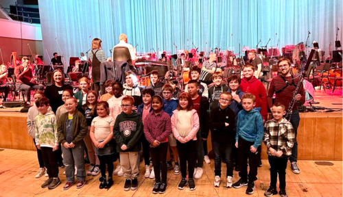 A group of children poses for a photo in front of an orchestra on stage, with musicians and instruments visible in the background.
