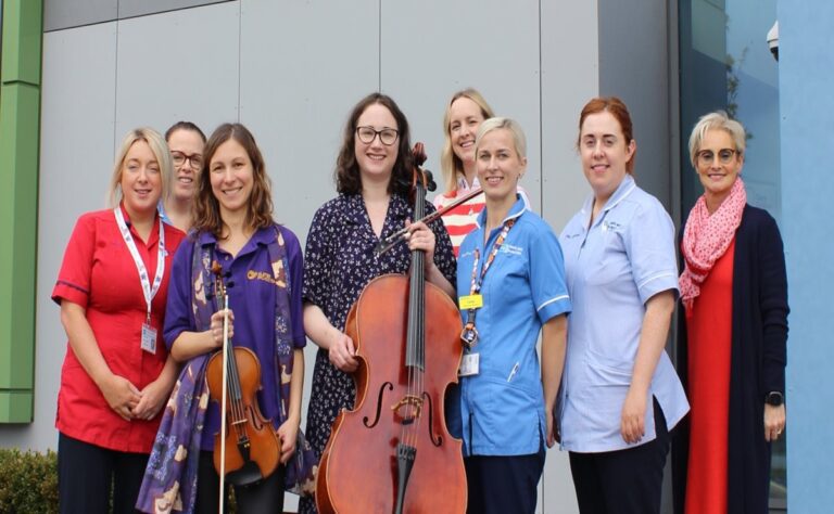 Eight women, including healthcare workers from Craigavon Area Hospital and two musicians with a violin and cello, stand together outside a modern building, highlighting the impact of music therapy on Blossom Children’s Ward.