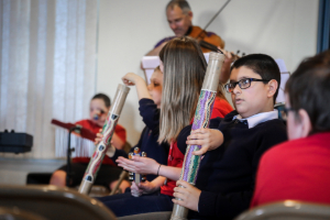 Children seated in a classroom hold decorated rain sticks, with a musician playing a violin in the background.
