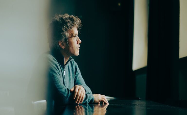 A man with curly hair and glasses sits at a table, looking thoughtfully out of a window in a dimly lit room, perhaps reflecting on Christoph Altstaedt’s recent Ulster Orchestra Good Friday concert.