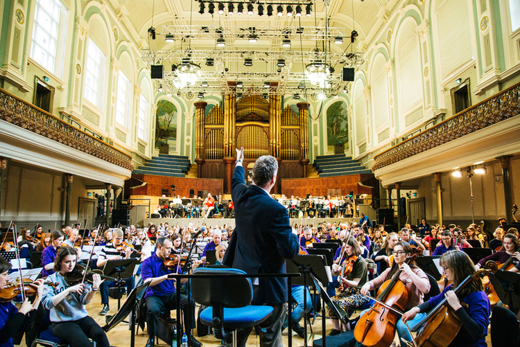 A conductor leads an orchestra rehearsal in a large, ornate concert hall as musicians play string instruments, their music filling the grand space.