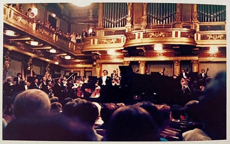 A symphony orchestra performs on stage in the grand, ornate Golden Hall of the Musikverein, with an audience seated in front and people watching from balconies, evoking the elegance of a 1986 concert night.