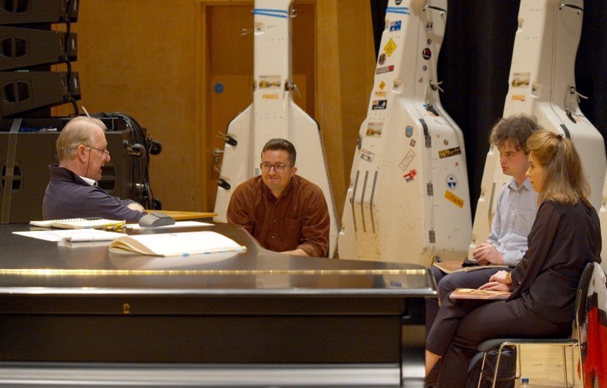 Four people sit around a piano in a rehearsal room, with several large instrument cases and music stands visible in the background.