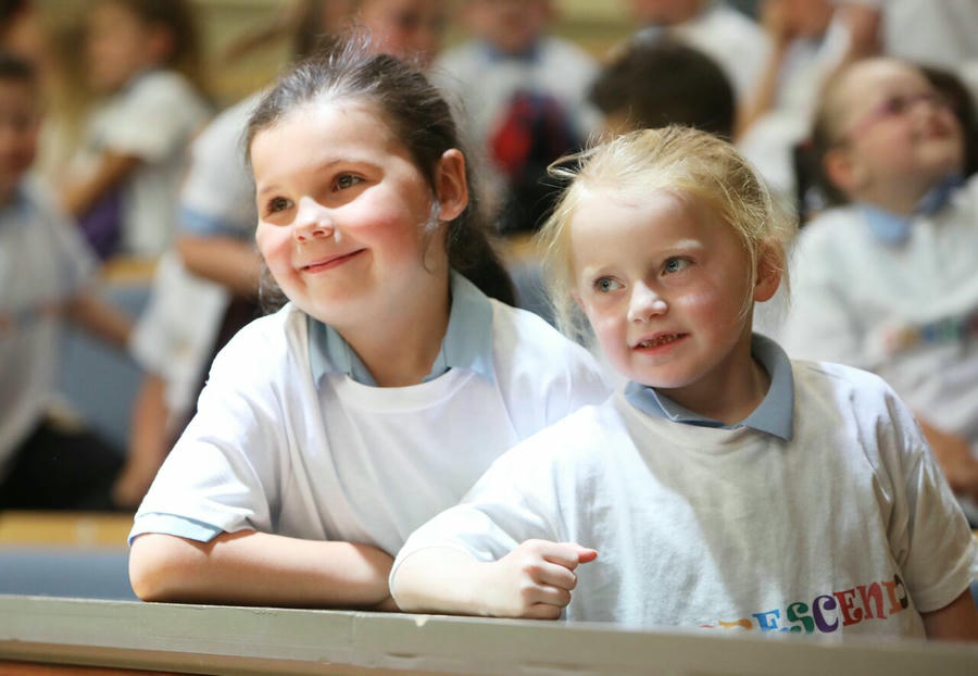 Two young girls wearing white t-shirts smile and look ahead while sitting together in what appears to be a school or group setting, with other children in the background.