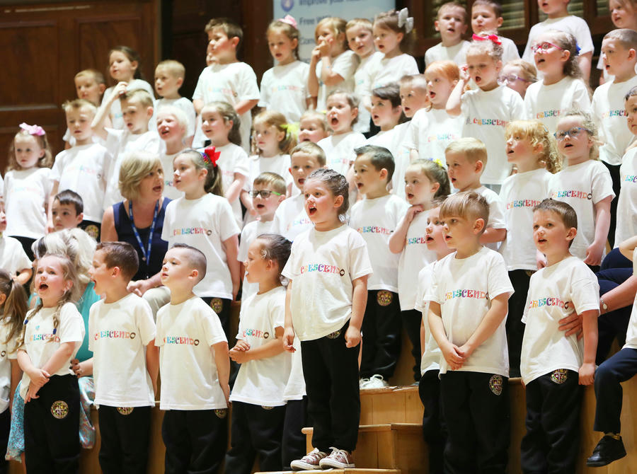 A large group of young children in matching white T-shirts sing together on stage with an adult seated among them.