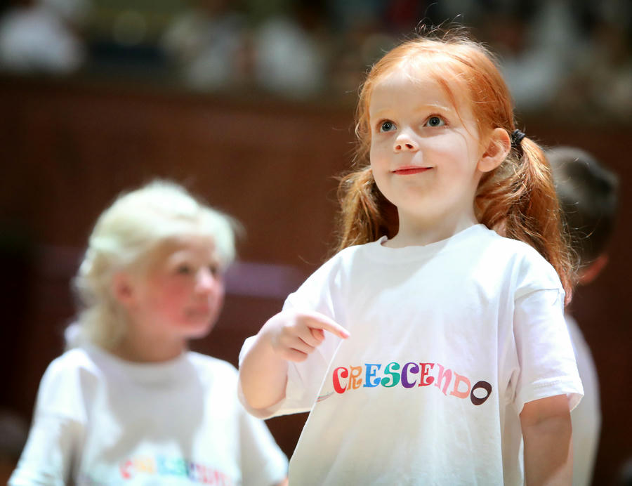 A young girl with red hair wearing a white "Crescendo" t-shirt stands indoors and points at her shirt. Another child blurred in the background.