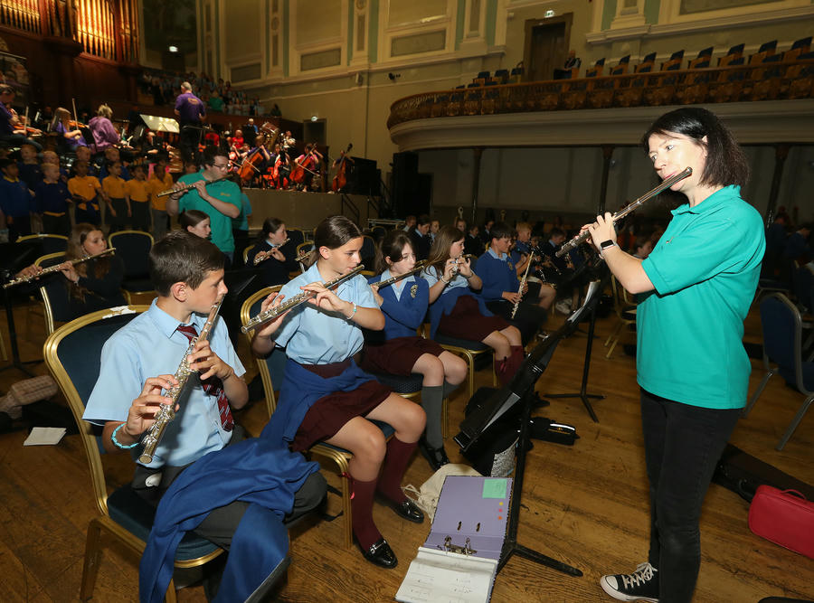 A music instructor guides a group of school students playing flutes in a concert hall, with an orchestra and choir visible in the background.