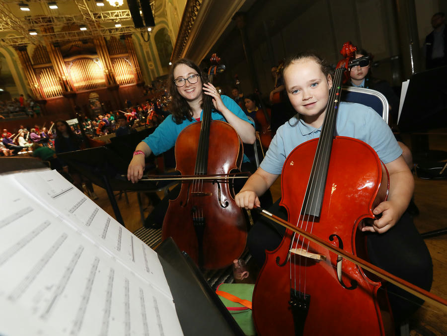 Two young girls seated and playing cellos in an orchestra setting, with sheet music in the foreground and other musicians in the background.