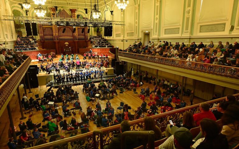 A large audience seated in a grand concert hall watches the orchestra and choir reach a stirring crescendo on stage; chandeliers glow above, with balcony seating framing this musical spectacle worthy of headlines.
