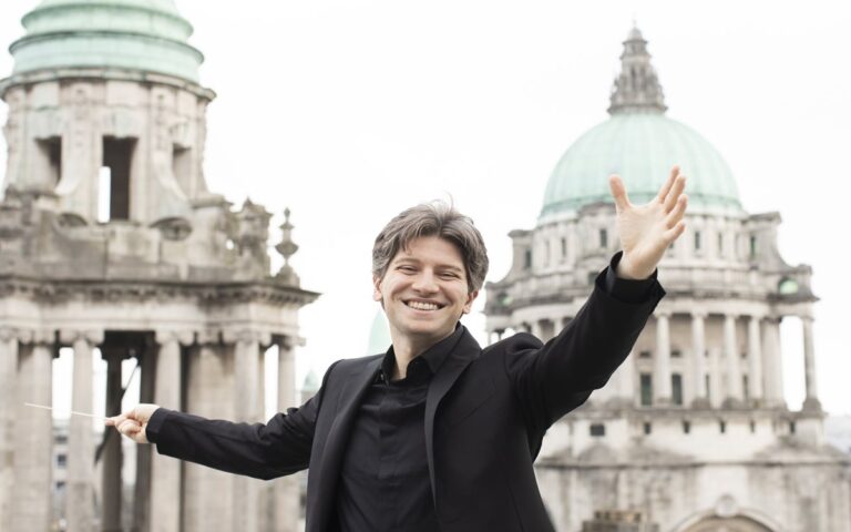 A smiling man in a black suit stands with arms outstretched, holding a conductor’s baton, in front of historic domed buildings.