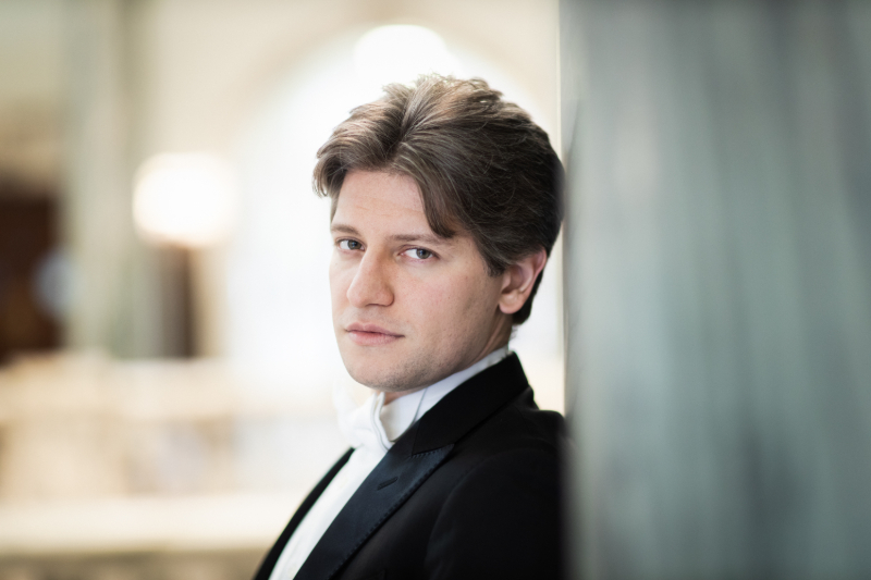 A man with light skin and brown hair, part of the Artistic Team, wears a black suit and white shirt as he leans against a marble wall in a softly lit indoor setting.