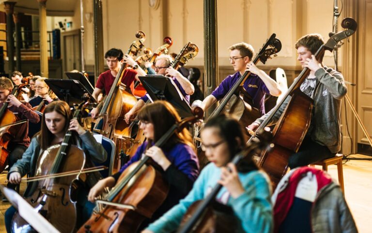 A group of musicians rehearses with cellos and double basses in a well-lit room, focused on their sheet music as an approaching deadline motivates them to perfect each note.