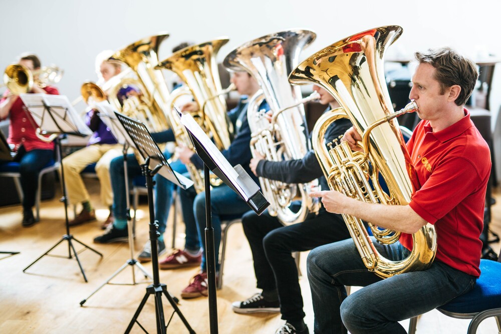 A group of people sitting in chairs play brass instruments, including tubas, in a well-lit room with music stands in front of them—a vibrant learning community captured for a community archive.