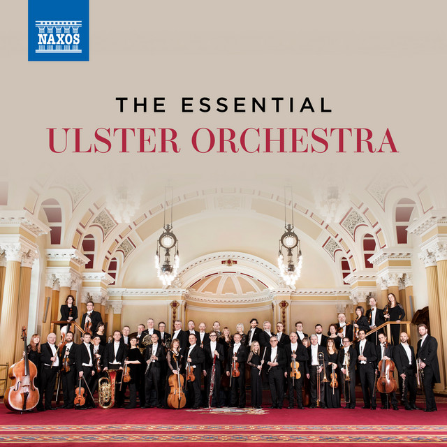 A large orchestra stands in formal attire holding instruments in an ornate hall under a sign that reads "The Essential Ulster Orchestra.