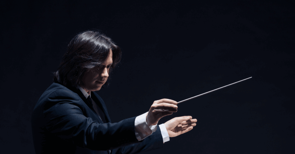A person in a black suit holds a conductor's baton and gestures with their hands against a dark background.