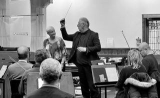 A conductor stands in front of seated musicians, holding a baton mid-air, while a woman stands beside him; music stands and sheet music highlight their involvement in collaborative projects.