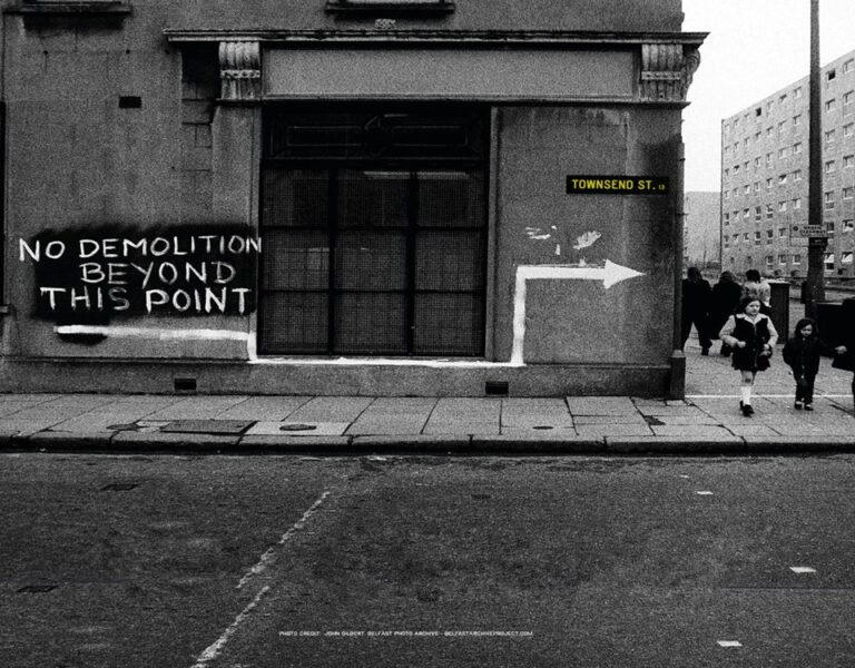 Black and white photo of Townsend Street’s corner, where a sign reads "NO DEMOLITION BEYOND THIS POINT," capturing both the heritage and future spirit as people walk along the sidewalk beside the building.