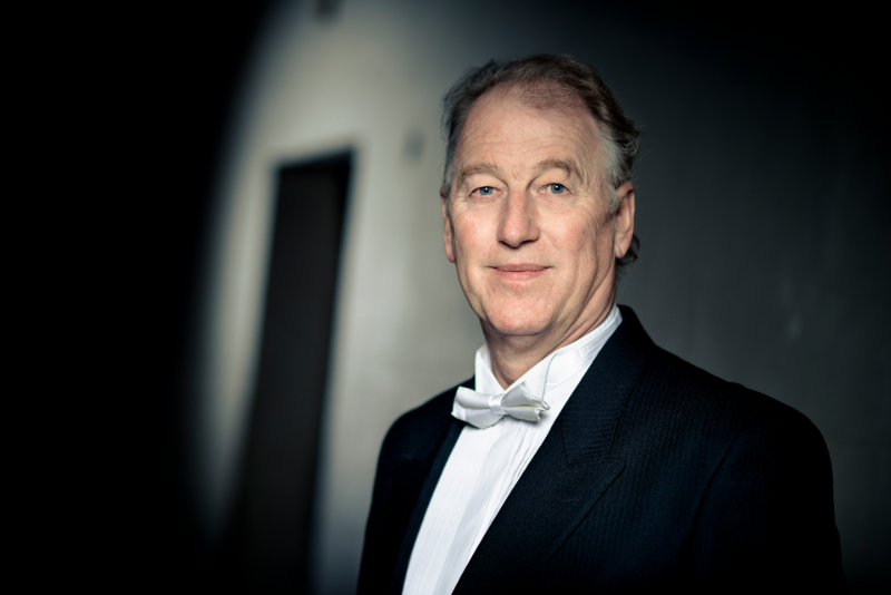 An older man in formal attire with a white bow tie and suit stands indoors, facing the camera and smiling slightly—an esteemed member of the Artistic Team.