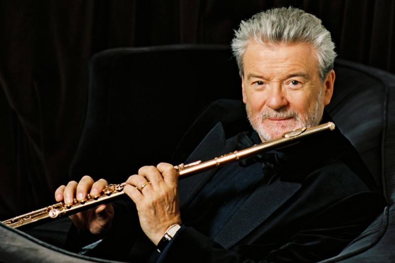 A gray-haired man in a black suit sits in a chair holding a flute, facing the camera with a slight smile, embodying the refined spirit of the Artistic Team.