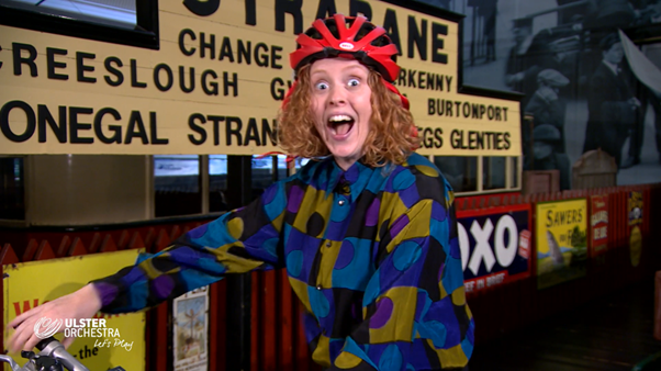 A person wearing a red bicycle helmet and colorful shirt smiles widely, standing next to a bicycle in front of vintage Irish railway signs and 2022 advertisements.