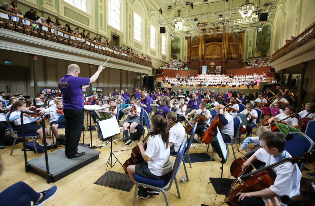 A conductor leads a youth orchestra and choir in a large concert hall, fostering a vibrant learning community as the audience enjoys the performance from the balcony.