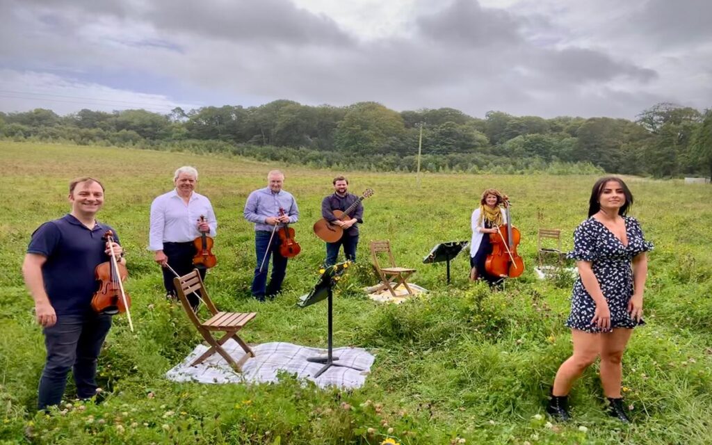 Six people with string instruments and a guitar perform in a grassy field with chairs and music stands under a cloudy sky, bringing the spirit of BBC Proms to life—it's like experiencing Proms in your garden.