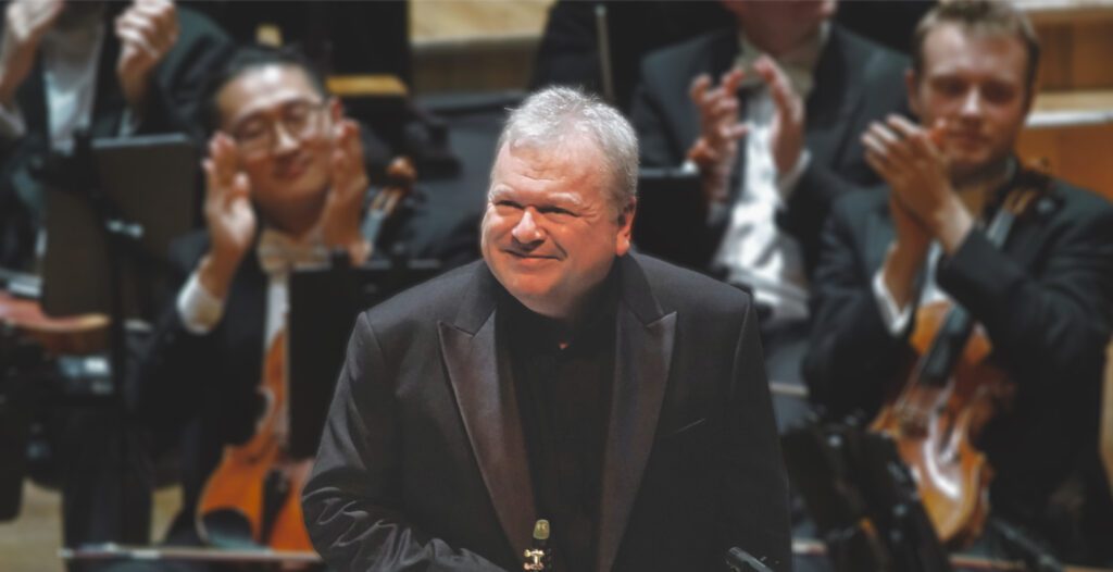 Principal Guest Artist Michael Collins stands in front of the Ulster Orchestra, smiling as musicians behind him applaud.